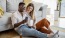 a man and woman with take out, sit against a couch on their living room floor and watch at their cell phone