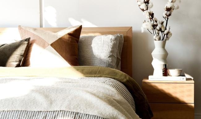 swath of sunlight shines across a well made bed and bedside table with a vase of cotton plants
